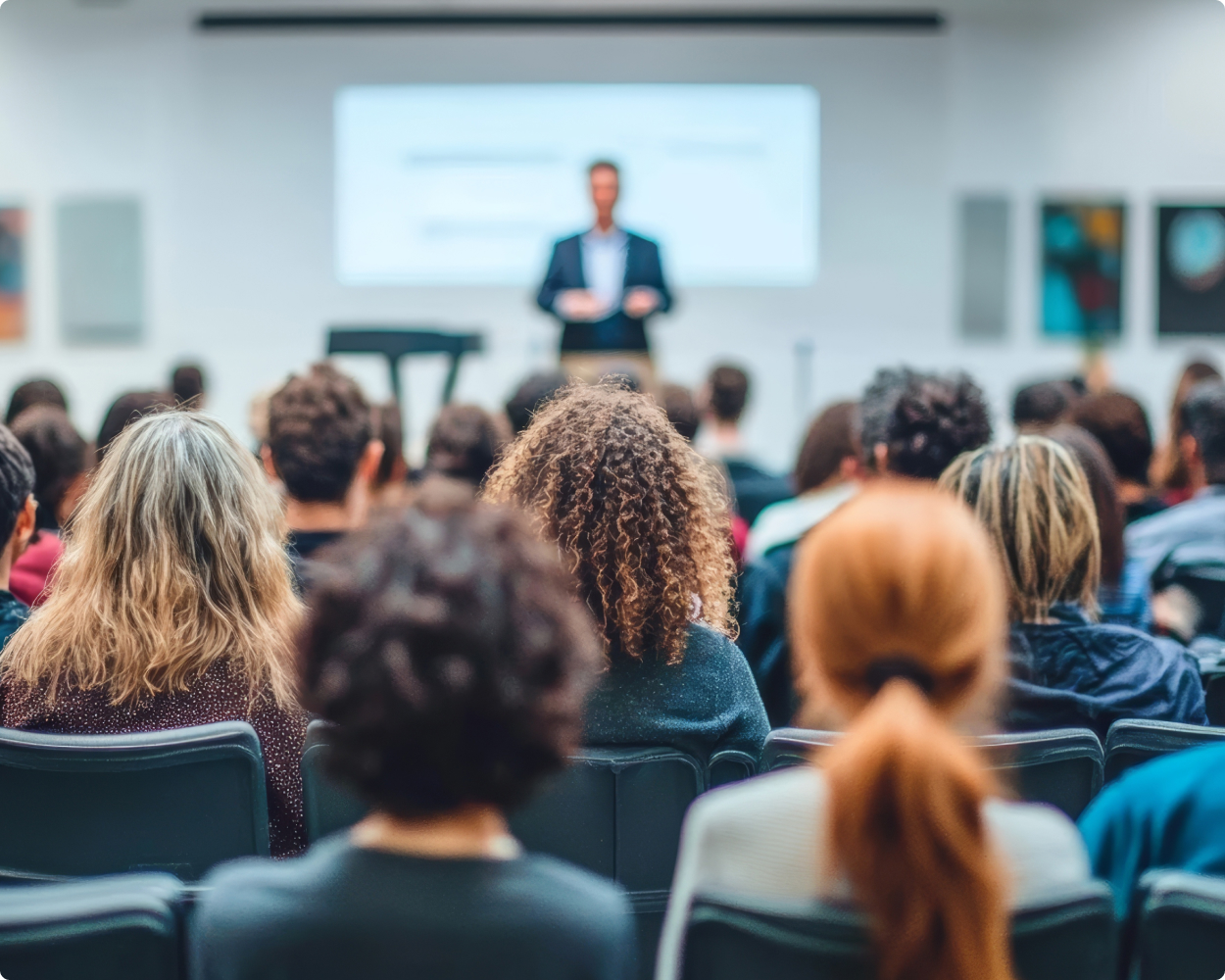 Ein Publikum lauscht einem Mann von aiwg, der vorne eine Präsentation hält und diese durch eine Power Point stützt.