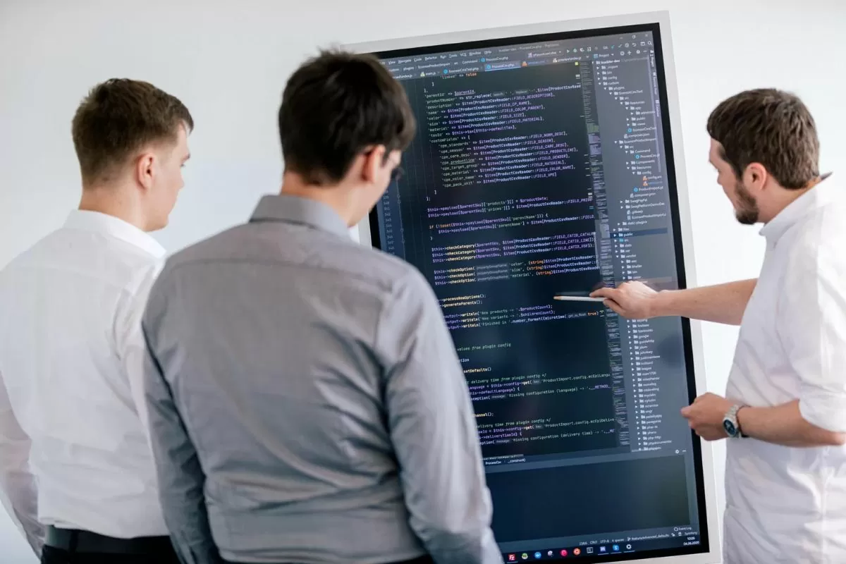 Drei Mitarbeitende stehen vor einem großen Bildschirm und gehen einen Code durch.