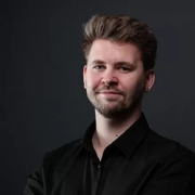 Ein Mann mit einem gepflegten Bart und einem schwarzen T-Shirt steht lächelnd da.