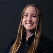 Eine Frau mit langen Haaren trägt ein schwarzes Shirt und schaut in die Kamera.