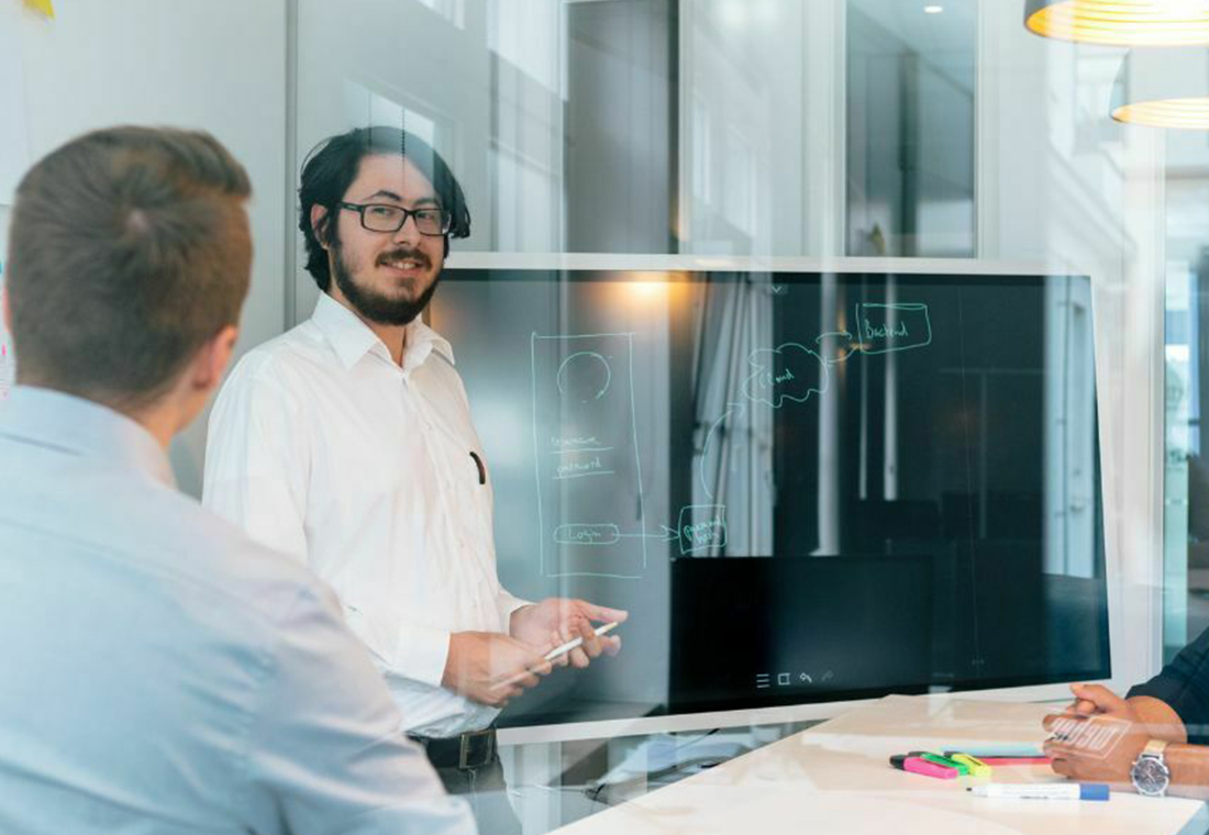 Eine Gruppe von Menschen in einem Besprechungsraum, die um einen Tisch versammelt sind, mit einem Whiteboard im Hintergrund.