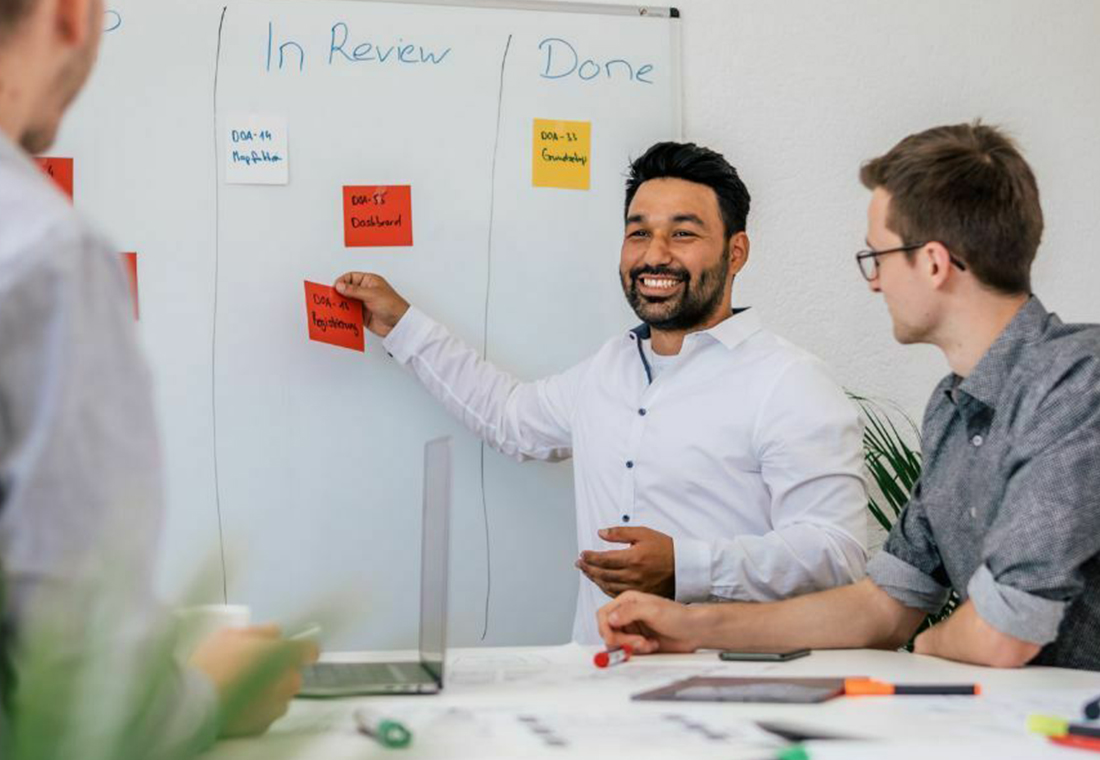 kategorietexte-online-shops Drei Männer stehen um ein Whiteboard mit bunten Haftnotizen und diskutieren Ideen.