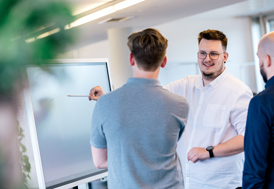 Drei Männer stehen vor einem Smartboard. Der Mann in der Mitte zeigt auf das Board.