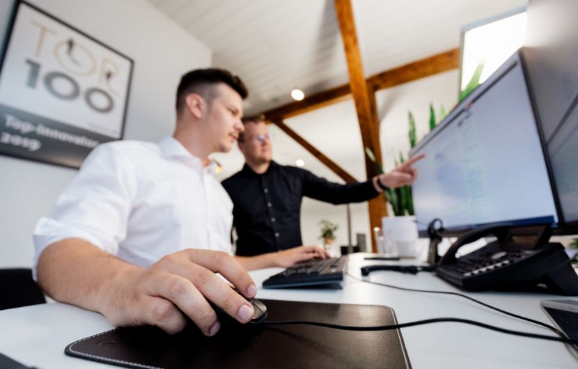 Zwei Mitarbeiter vor einem Bildschirm, einer der beiden zeigt dem anderen etwas auf diesem