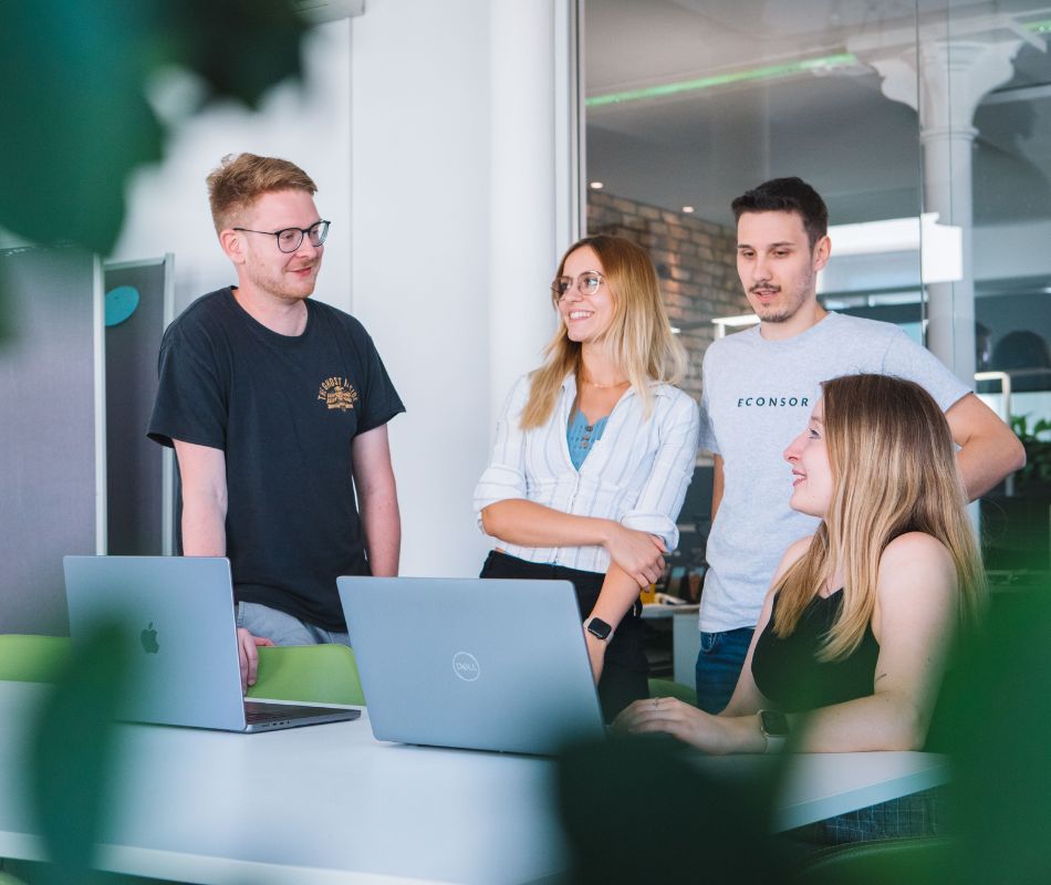 Vier Mitarbeiter sind im Besprechungszimmer und unterhalten sich  vor zwei Laptops