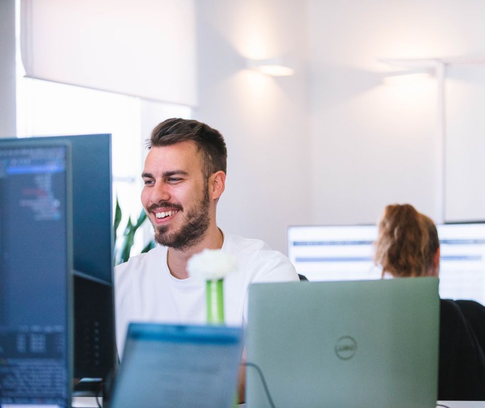 Mitarbeiter lächelt, sitzt am Schreibtisch und schaut auf seinen Bildschirm.