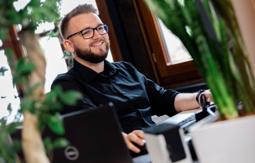 Laechelnder-Geschaeftsmann-sitzt-vor-Computer-und-arbeitet-Vordergrund-eine-Pflanze. Lächelnder Geschäftsmann sitzt vor Computer und arbeitet, Vordergrund eine Pflanze.