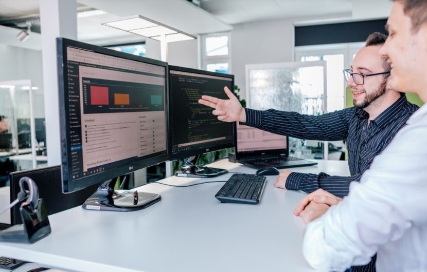 Zwei-Geschaeftsleute-sitzen-an-Tisch-und-gemeinsam-schauen-in-Computer-Geschaeftsmann-zeigt-mit-Hand-auf-Bildschirm. Zwei Geschäftsleute sitzen an Tisch und gemeinsam schauen in Computer Geschäftsmann zeigt mit Hand auf Bildschirm.