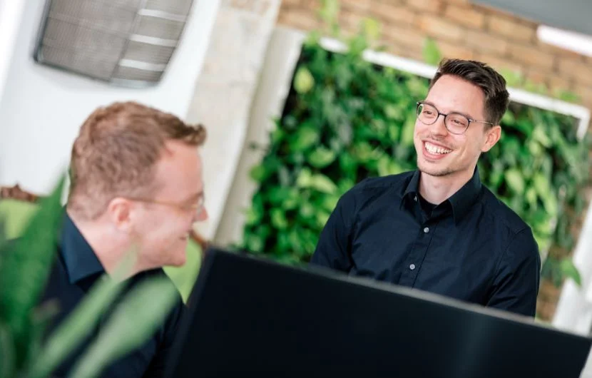 Agentur für Onlineshops Zwei Online-Shop-Experten in einem modernen Büro mit begrüntem Hintergrund, die sich lächelnd über Suchmaschinenoptimierung unterhalten.