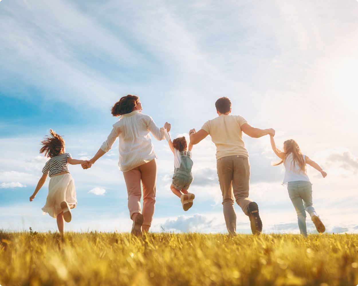 Eine Familie von fünf, die bei BKK Verbund Plus versichert ist, läuft händchenhaltend über eine Wiese an einem Sommertag.