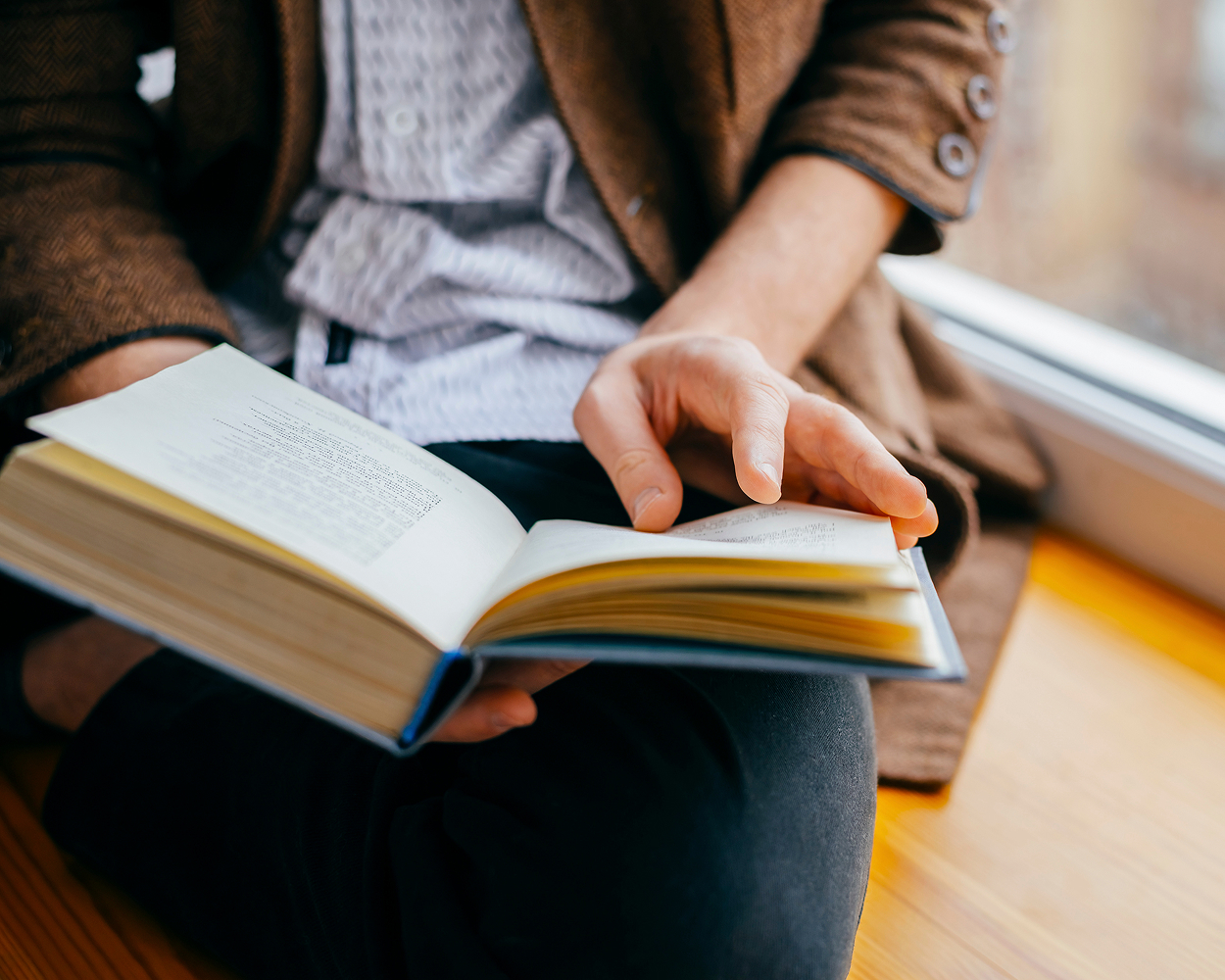 Ein Mensch, der auf einer Holzbank am Fenster sitzt und ein Buch vom Carl Auer Verlag in der Hand hält und blättert