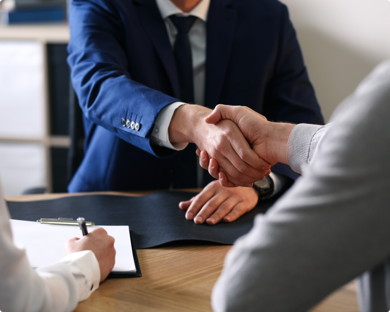 Zwei Männer in einem Anzug schütteln sich die Hände, während eine dritte Person, ein Anwalt von db-anwälte etwas auf einem Papier notiert