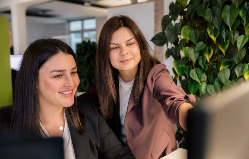 Zwei Kolleginnen arbeiten zusammen an einem Computer und betrachten den Bildschirm, während sie eine Lösung für ein Portal entwickeln.