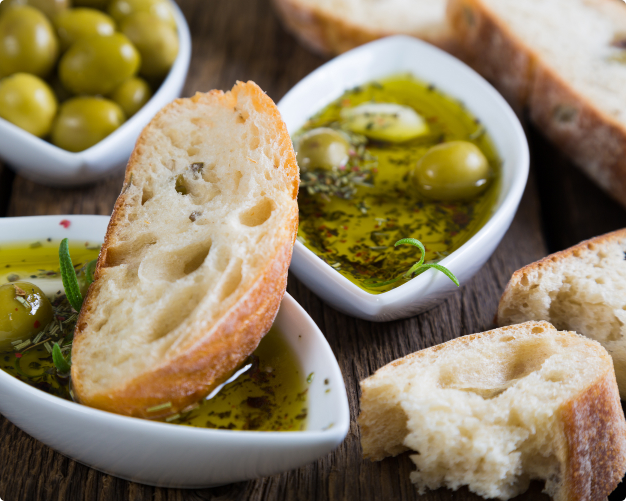 Schälchen mit Oliven und Olivenöl von edesma liegen auf einem Holztisch. Daneben liegen noch ein paar geschnittene Scheiben Brot und eine Scheibe davon liegt in einem Oolivenölschälchen.