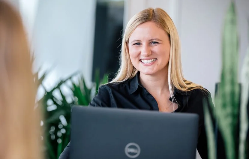 Lächelnde Frau mit blondem Haar sitzt an einem Schreibtisch hinter einem Laptop, umgeben von grünen Pflanzen – sie wirkt freundlich und professionell.