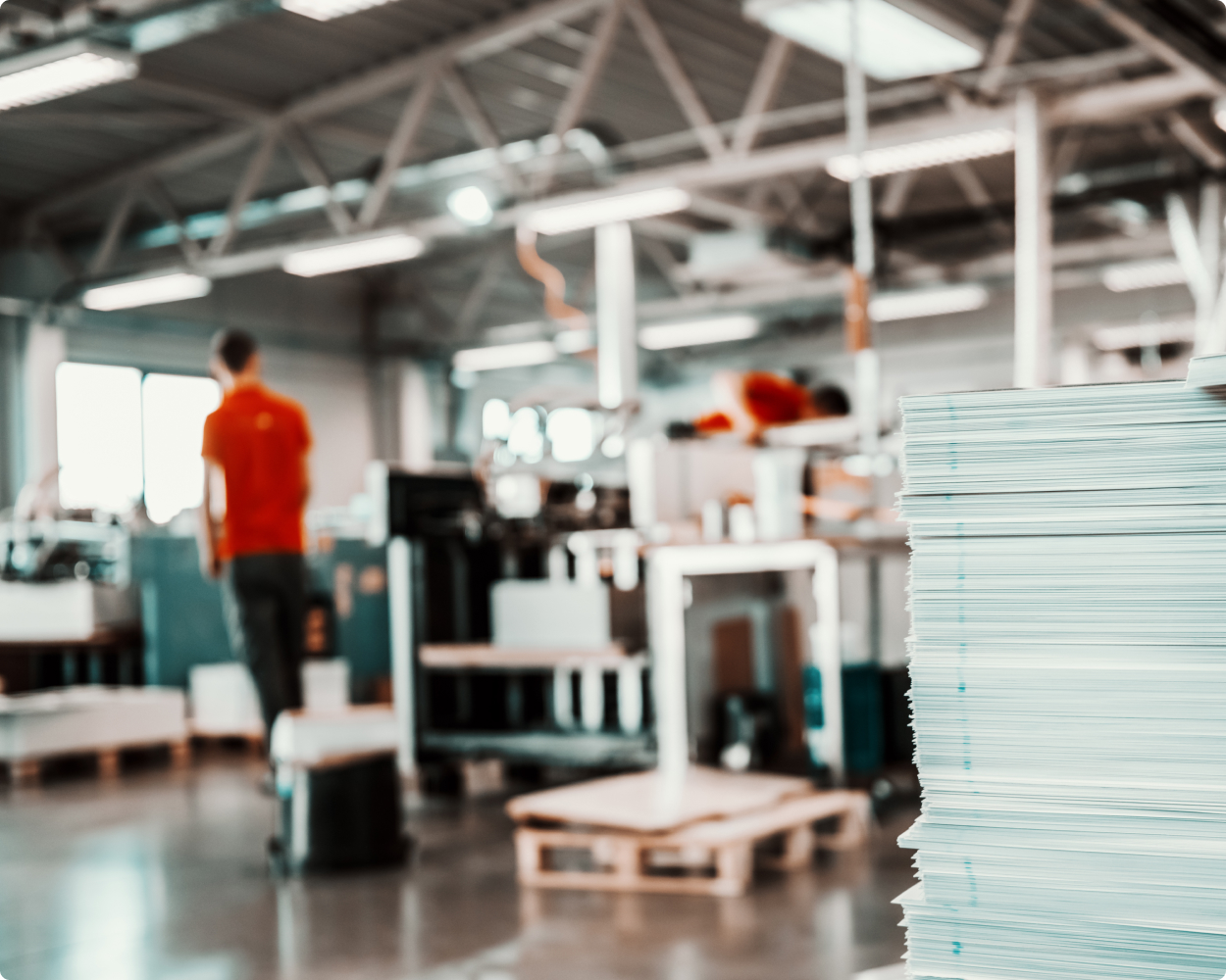 Stapel von Dokumenten im Vordergrund einer ISIMAT-Produktionshalle, im Hintergrund unscharf Mitarbeitende bei der Arbeit – Fokus auf strukturierte Fertigungsprozesse.