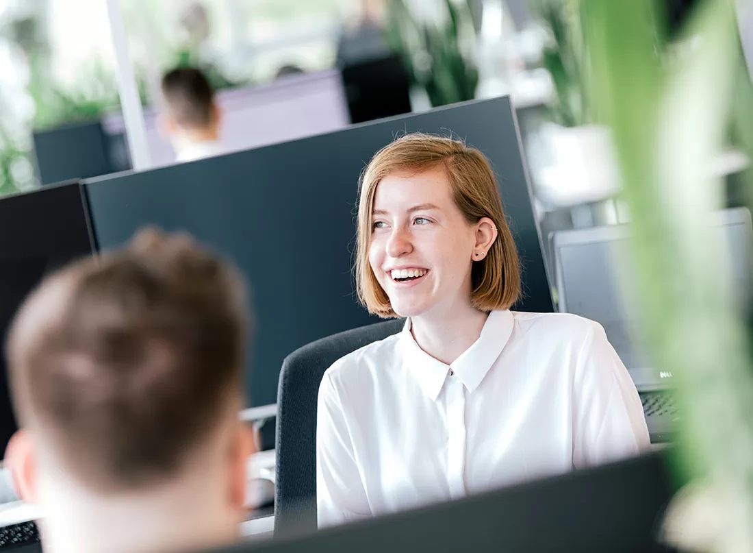 Eine Mitarbeitende sitzt im Büro und lächelt einen Kollegen an