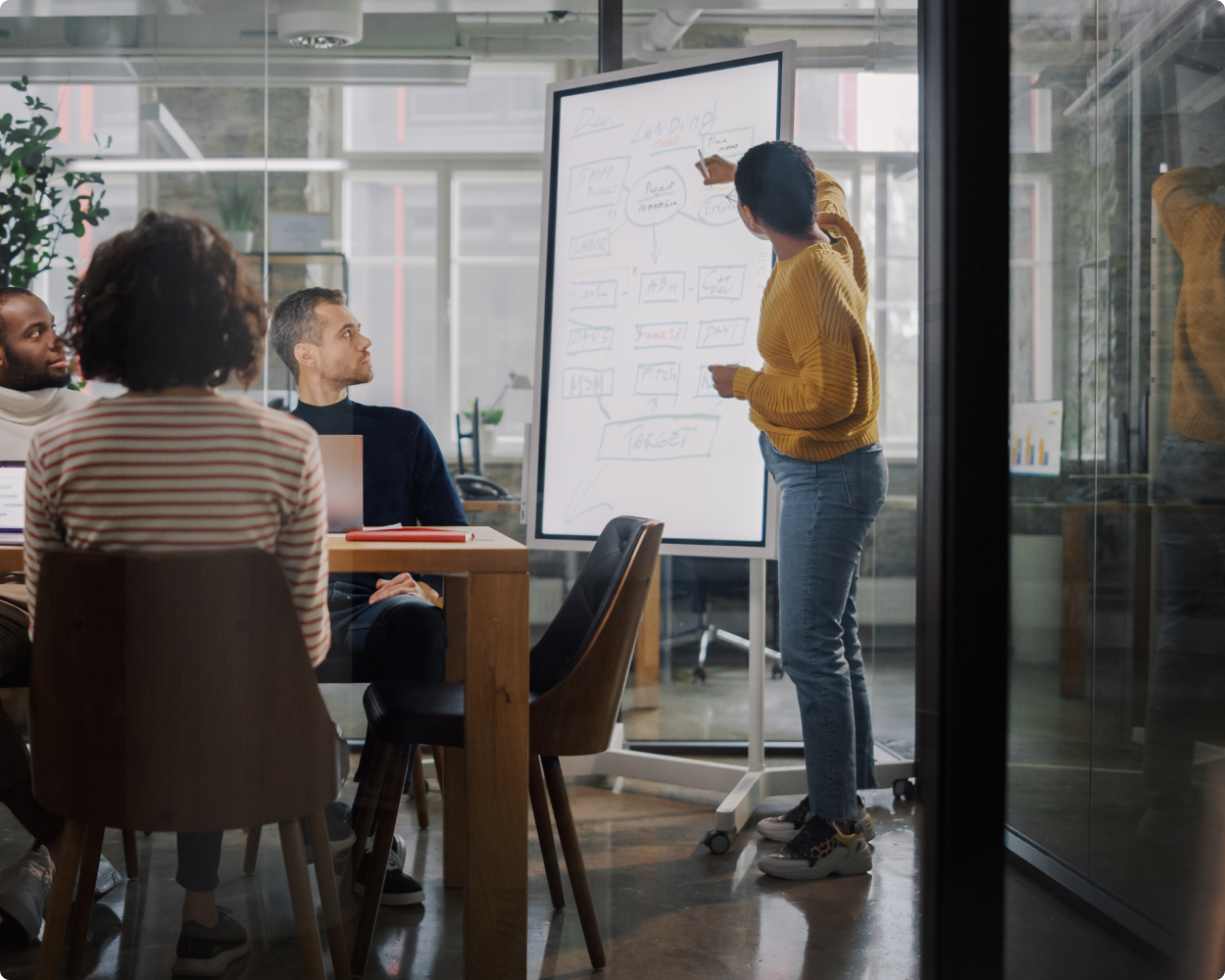 Eine Person präsentiert eine Strategie an einem Whiteboard vor einem kleinen Team in einem modernen Konferenzraum – kreativer Austausch bei JOM Consulting.