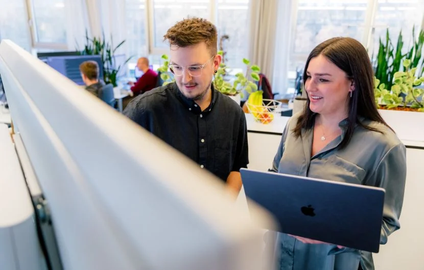 Zwei Mitarbeiter stehen vor einem Whiteboard, während einer der beiden etwas präsentiert.