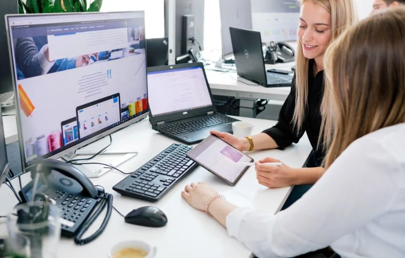 Ihre Online-Marketing-Agentur in Köln Zwei Mitarbeiterinnen sitzen am Schreibtisch während eine der beiden etwa auf einem Tablet zeigt.