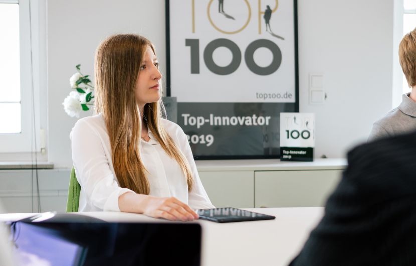 Junge Frau in einer modernen Online-Marketing-Agentur sitzt am Tisch mit Laptop; im Hintergrund erhellt ein 'Top 100 – Top-Innovator 2019'-Schild die professionelle Arbeitsatmosphäre.