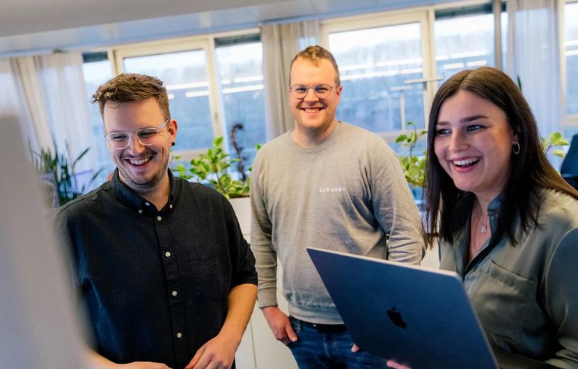 Ihre Performance-Marketing-Agentur in Stuttgart Drei Teammitglieder von ECONSOR lachen gemeinsam, während sie auf einen Laptop schauen und sich über ein Projekt austauschen. Im Hintergrund sind Pflanzen und ein Büro mit großen Fenstern zu sehen.