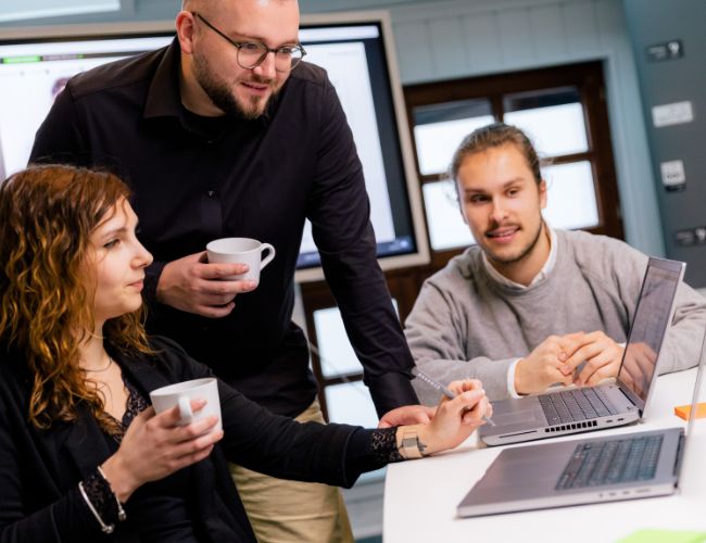Ihre Agentur für E-Commerce und Portale Ein Team aus drei Personen steht zusammen und schaut auf einen Laptop, während sie eine Besprechung zu E-Commerce und Portalentwicklung führen.