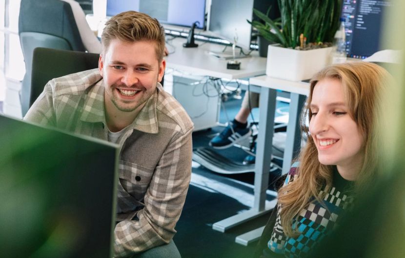 Zwei Personen arbeiten zusammen an einem Computer, lächeln und haben eine angenehme Besprechung in einem modernen Büro. Im Hintergrund sind weitere Arbeitsplätze sichtbar.