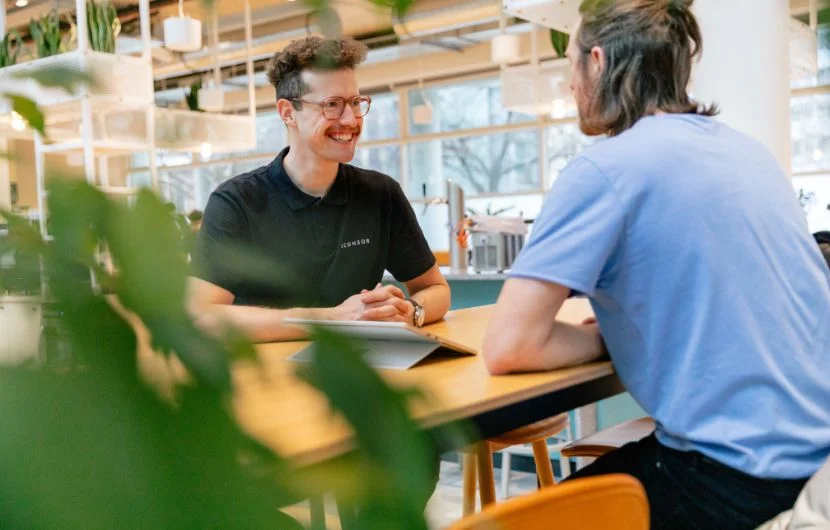 Ihre SEO-Agentur in München Zwei Mitarbeiter, einer davon lächelt, der andere steht mit dem Rücken zur Kamera sitzen an einem Tisch und unterhalten sich.