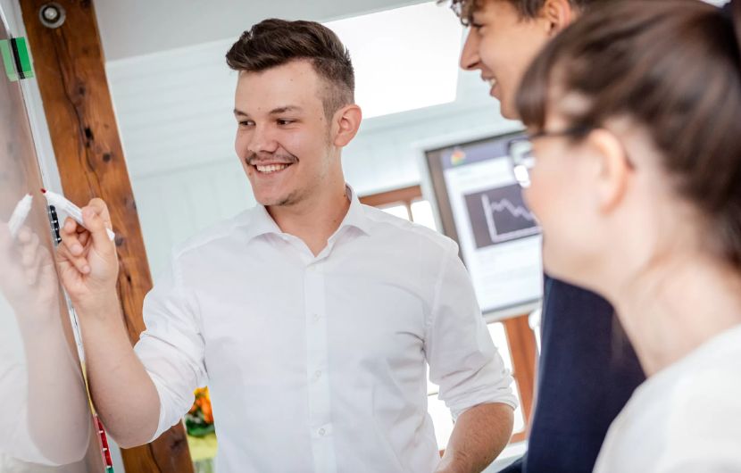 Ihre Social-Media-Agentur für zielgerichtetes Marketing Ein junger Mann in weißem Hemd erstellt zusammen mit zwei Kolleg*innen eine Social-Media-Strategie an einem Whiteboard – symbolisiert die Zusammenarbeit in einer Social-Media-Agentur, die auf effektives Social-Media-Marketing setzt.