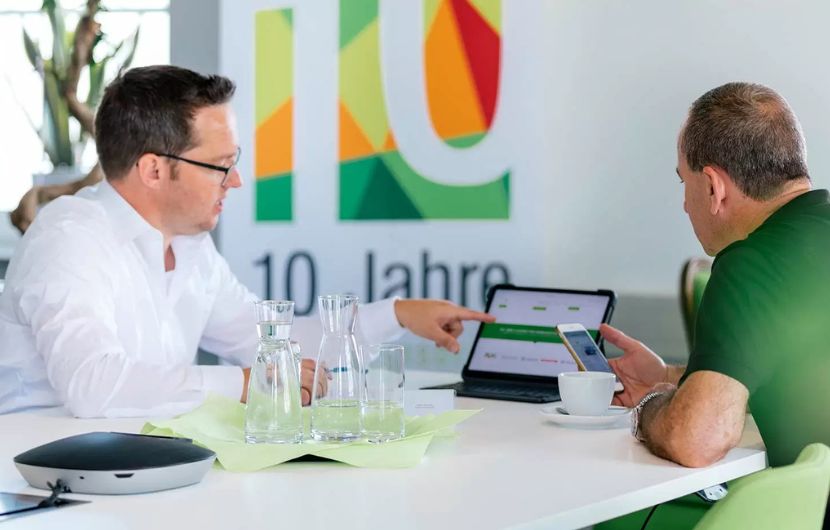 Ein Systemberater spricht mit einem Kunden über eine technische Lösung. Beide sitzen an einem Tisch in einem modernen Büro mit Pflanzen im Hintergrund.