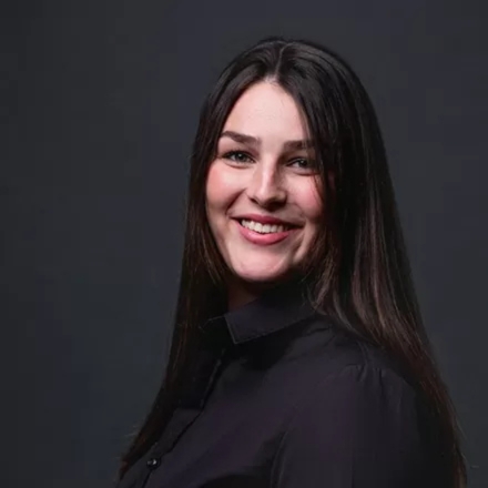 Eine Frau mit langen Haaren trägt ein schwarzes Shirt und blickt in die Kamera.