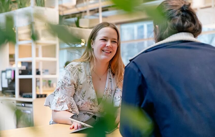 webdesign-agentur-muenchen Zwei Mitarbeiter sitzen an einem Tisch und unterhalten sich. Die Mitarbeiterin lächelt und hält ein Tablet in der Hand.