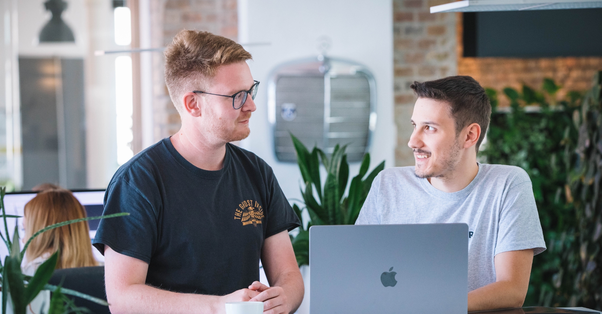 Zwei Kollegen im Austausch am Arbeitsplatz, auf dem Tisch stehen ein Laptop und eine Tasse Kaffee.