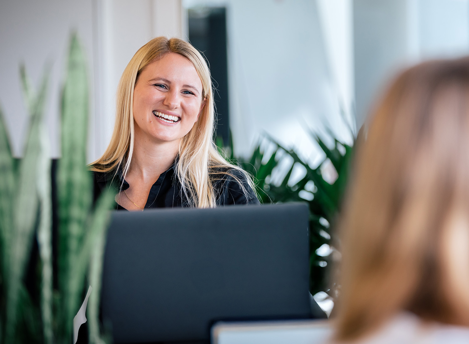 Lächelnde Mitarbeiterin der Digitalagentur ECONSOR im Gespräch mit Kollegin, aufgenommen in moderner Büro-Umgebung mit Pflanzen im Vorder- und Hintergrund.